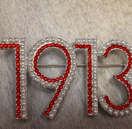Brooch with '1913' design featuring red stones and pearls on a textured background