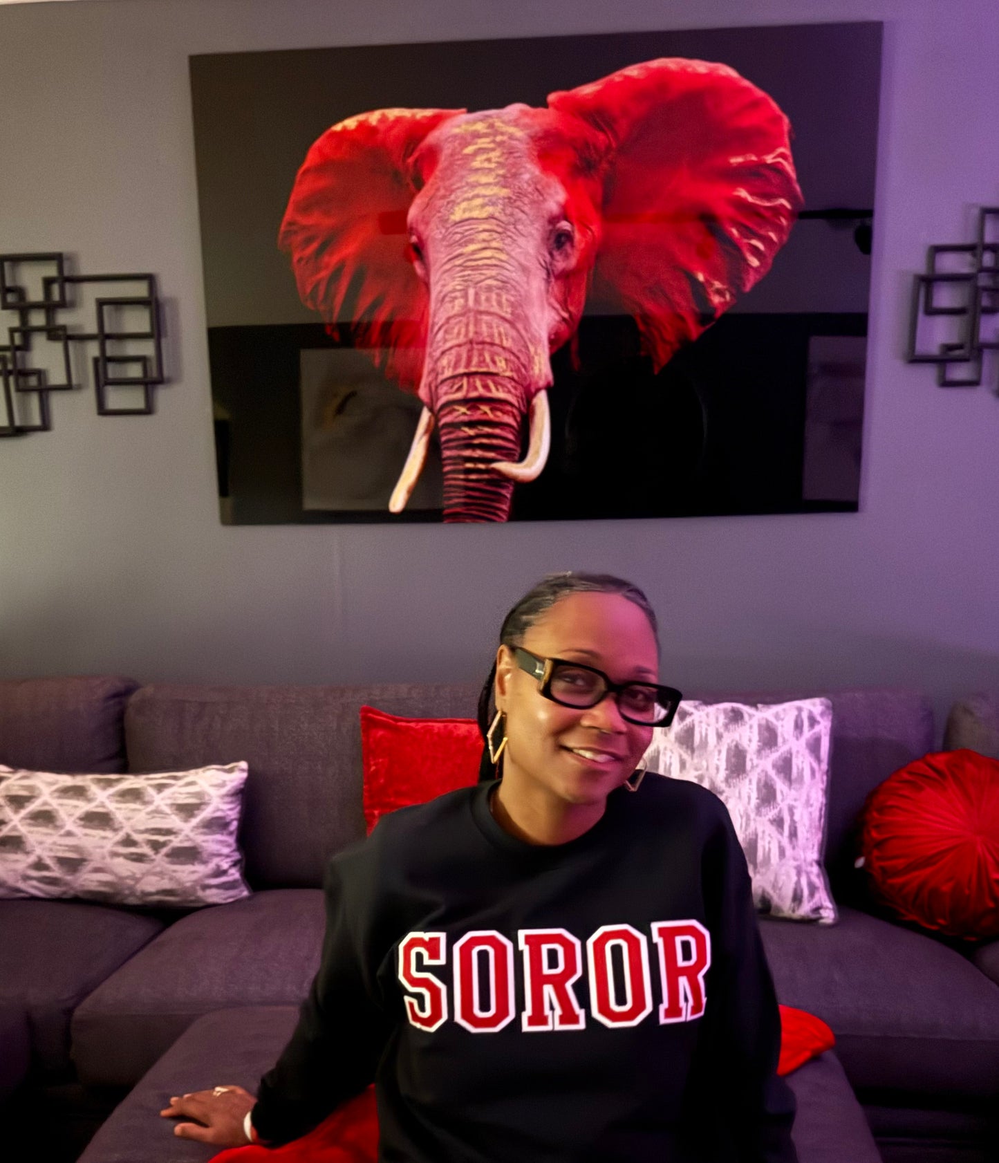 Woman wearing a black sweatshirt with 'SOROR' in red letters, sitting on a couch in a room with a large elephant painting on the wall.