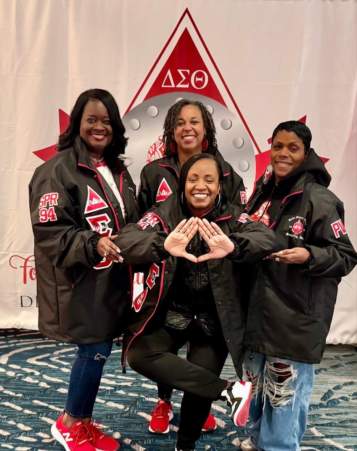 Four women in matching jackets posing in front of a Delta Sigma Theta banner.